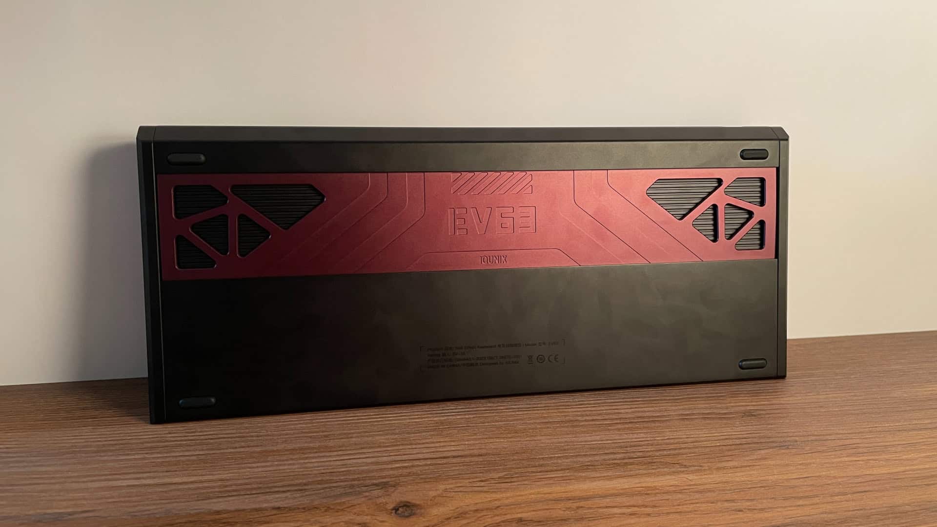 Bottom view of the IQUNIX EV63 mechanical keyboard showing a decorative weight in matching burgundy finish, four elongated rubber feet for stability, and industrial-style design elements that continue the cohesive aesthetic from the rest of the keyboard.
