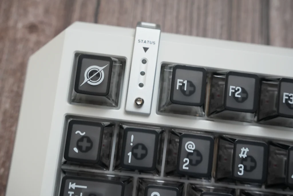 Close-up of the upper-left corner of a white MelGeek keyboard. A slim metal STATUS strip sits between keys, marked with a downward triangle and three indicator dots. Beside it is a dark keycap with a white circle-and-slash emblem, then translucent F1 and F2 keycaps.
